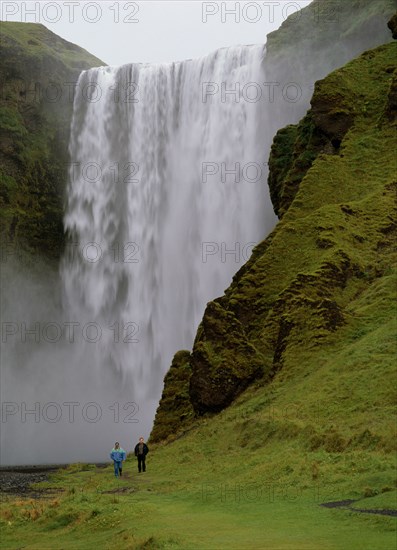 ICELAND, Skogafoss Waterfall, Cascading waterfall 60 metres or 200 feet high.  Two people walking along footpath at side.