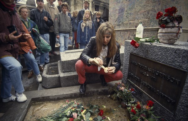 FRANCE, Ile de France, Paris, Pere Lachaise Cemetery. Visitors surrounding Jim Morrison grave with a woman knelling by the grave stone lighting a candle