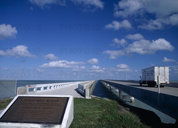 USA, Florida Keys, Seven Mile Bridge. Commemorative plaque with the Honorable Florida State Representative Bernie C Papy seen in the foreground with a truck driving along the concrete bridge stretching out over the sea