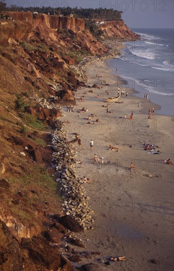 INDIA, Kerala, Varkala, "Beach sheltered by steep, eroded cliffs with sunbathers on the sand and palm trees on headland beyond."