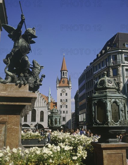 GERMANY, Bavaria, Munich, Marianplatz with people walking below the clock tower with a stone plinth and flower boxes in the  foreground