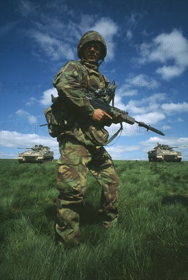 MILITARY, Soldier, Training, Soldier in camouflage standing in a field holding a gun with two Warrior tanks behind