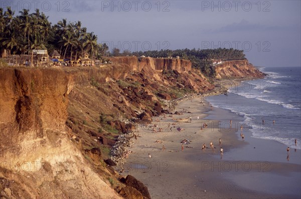 INDIA, Kerala, Varkala , "Coastline and narrow sandy cove with sunbathers sheltered by steep, eroded cliffs with bars and restaurant backed by palm trees along top."