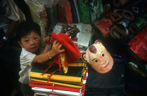 CAMBODIA, Phnom Penh, Central Market.  Small boy in shop selling fabric and flags. Coloured mask beside him.