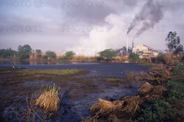 INDIA, Uttar Pradesh , Sugar Cane, Sugar mill emitting dark clouds of smoke from chimneys beside polluted lake.