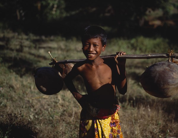 INDONESIA, Lombok, Timur, Mawon near Kuta a young boy in floral sarong holding pole of dried coconuts across his shoulder