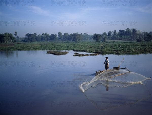 INDONESIA, Lombok, Timur NTB, "Praya, man fishing from outrigger canoe using circular casting net, lake weeds & trees behind J6599 "