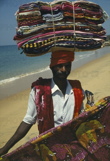 INDIA, Kerala, Kovalam Beach, Fabric salesman on beach carrying stack of colourful materials on his head.