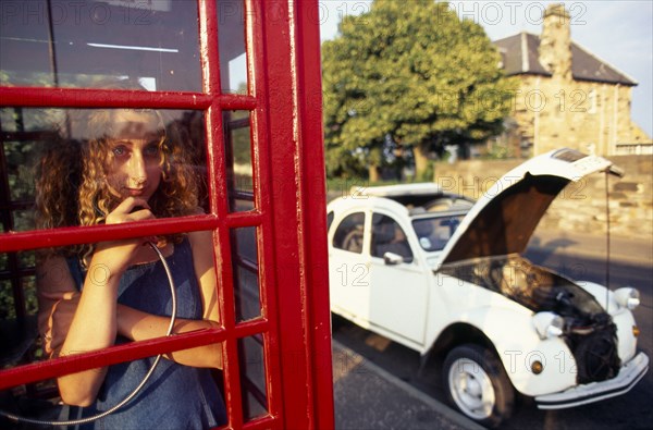 COMMUNICATIONS, Telephone, Phone Box, Girl making a call in a traditional red phone box with broken down Citroen 2CV car outside