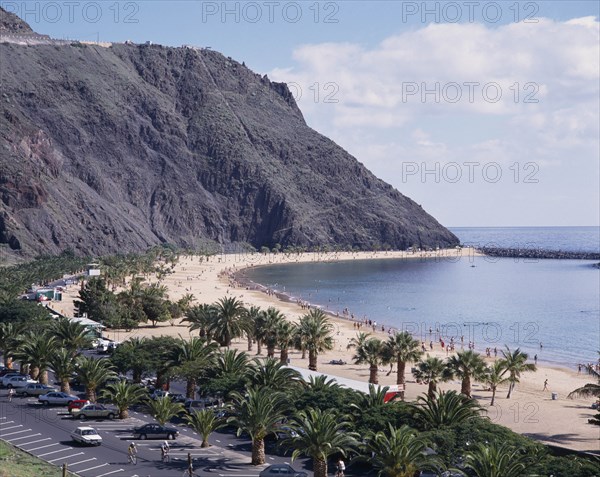 SPAIN, Canary Islands, Tenerife, Las Teresitas. Long sandy bay beneath black cliffs and palm lined promenade with parked cars