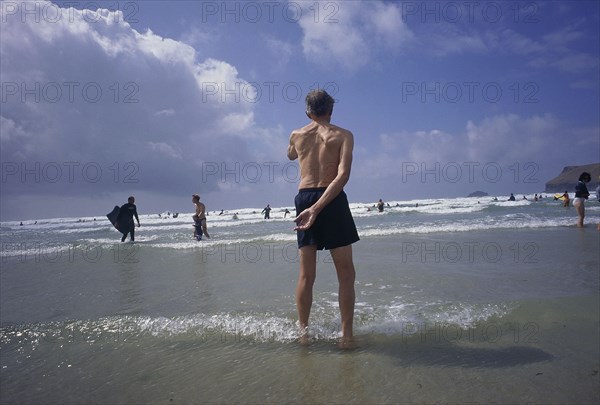 ENGLAND, Cornwall, Polzeath, Man standing in shallow water on the beach looking out to bathers in the sea