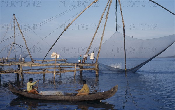 INDIA, Kerala, Kochi, Chinese fishing net being raised with a catch as two men row past in a canoe and a white ship sits on the horizon