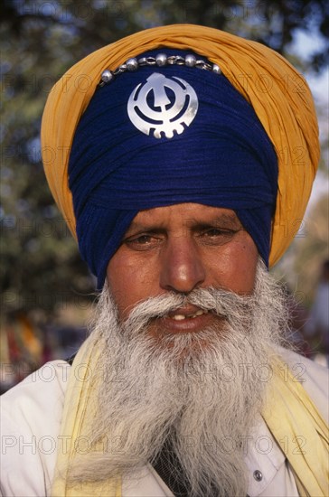 INDIA, Delhi, "Portrait of a sikh man wearing orange and royal blue  turban set with a khanda, the symbol of Sikhdom and with grey beard and moustache."