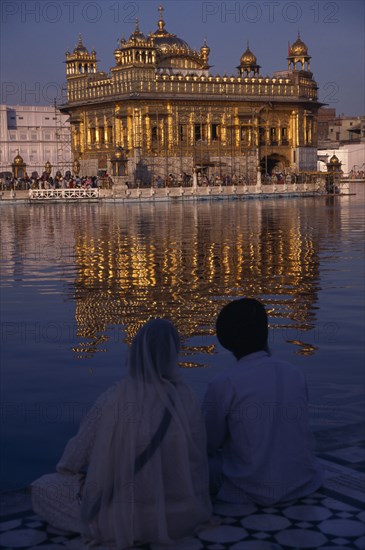 INDIA, Punjab, Amritsar, Sikh couple sitting on marble walkway looking out across sacred pool towards the Golden Temple and shimmering reflection on rippled water surface.