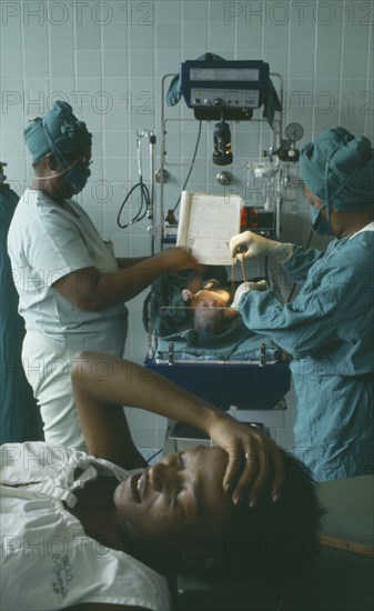 CUBA, Havana, Woman lying with her hand on her head after giving birth while two nurses weigh her baby behind