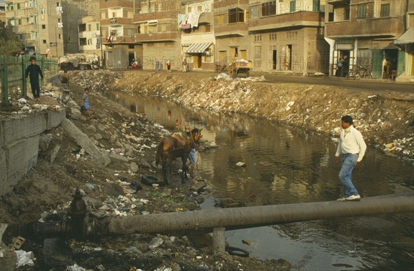 EGYPT, Damietta, Nile Delta, Rubbish in canal with man walking over pipe