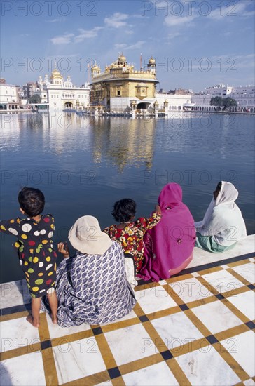 INDIA, Punjab, Amritsar , Women and children sitting on walkway beside sacred pool looking across to the Golden temple reflected in rippled surface of water.
