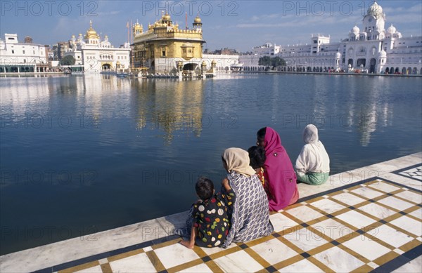 INDIA, Punjab, Amritsar, Three women and children sitting on marble walkway beside the sacred pool looking towards The Golden Temple.