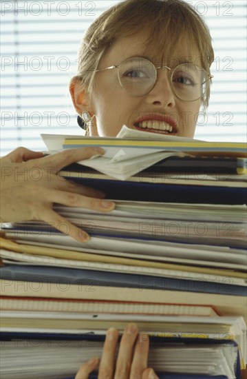 BUSINESS, Paperwork, Female office worker with stressed expression weighed down with stack of paperwork.