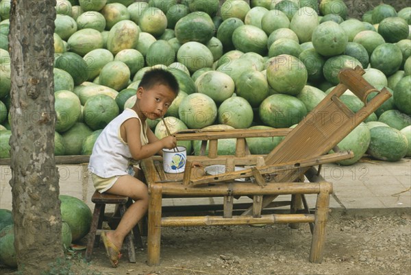 CHINA, Guizhou Province, Kaili, Liitle boy eating from bowl with chopsticks with large pile of watermelons behind.