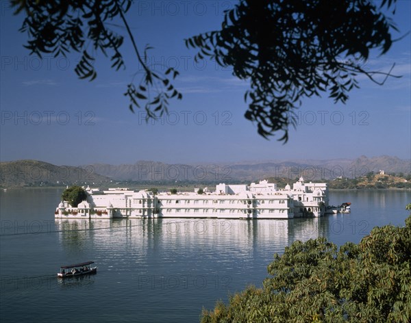 INDIA, Rajasthan, Udaipur, The Lake Palace framed by tree branches with white reflections on water and a boat travelling past