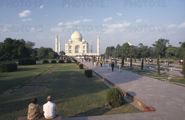 INDIA, Uttar Pradesh, Agra, Taj Mahal and gardens with couple sitting in the foreground and other people on a path leading up to the building
