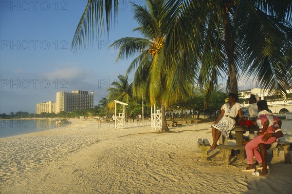 WEST INDIES, Jamaica , Ocho Rios , Mallard beach with people sitting beneath palm tree in foreground and hotel beyond.