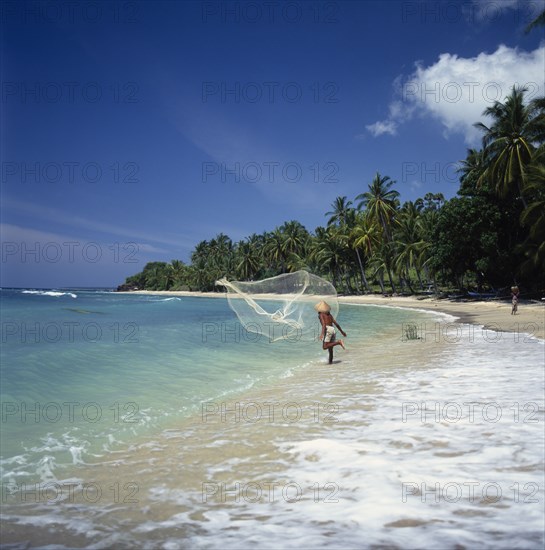 INDONESIA, Lombok, Mentiggi, Fisherman casting fishing net into sea from the surf on a coconut palm tree lined sandy beach