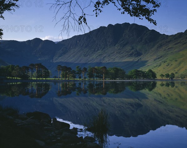 ENGLAND, Cumbria, Lake District, Buttermere.  Rugged hills and trees with mirror reflection in lake.
