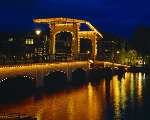 HOLLAND, North, Amsterdam, Magere Brug or Skinny Bridge illuminated at night with lights reflected in the canal below.