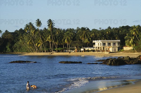 WEST INDIES, Tobago, Grafton Beach, People standing in sea beside sandy beach with white painted building surrounded by palm trees.