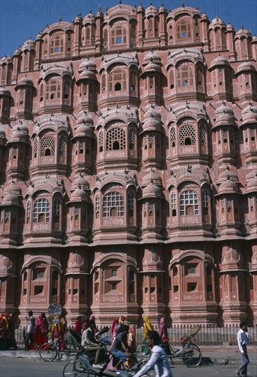 INDIA, Rajasthan, Jaipur, "Hawa Mahal or Palace of the Winds.  Part view of semi-octagonal, honeycombed sandstone facade.   Cyclists and brightly dressed women on street in front. "