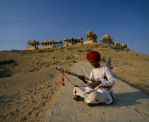 INDIA, Rajasthan, Jaisalmer, Musician wearing a red turban sitting cross-legged on the ground with old buildings on top of the hill behind him