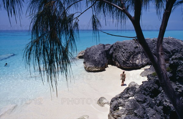 BERMUDA, Whale Bone Bay Beach, Female tourist in dress walking along the beach towards rocks seen through trees. Clear turquoise sea.