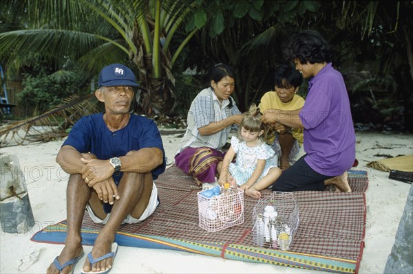 THAILAND, South, Koh Samui, Young Western girl on sandy beach having her hair braided by three Thai women with man sitting smoking in front and palm trees behind.