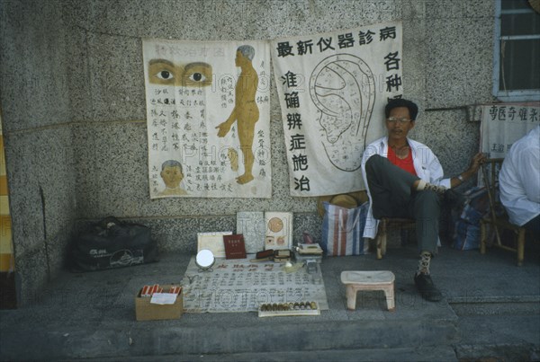 CHINA, Hainan Island, Sanya, Roadside medicine seller seated in front of medical drawings of the human body