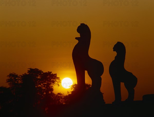 CAMBODIA, Angkor, Angkor Wat.  Golden setting sun beyond silhouetted trees and statues.