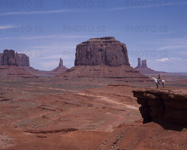 USA, Arizona, Monument Valley, Mittens desert rock features. Isolated blocks and pinnacles and flat valley floor with road running through. Rider on horse admires view from rocky ledge.