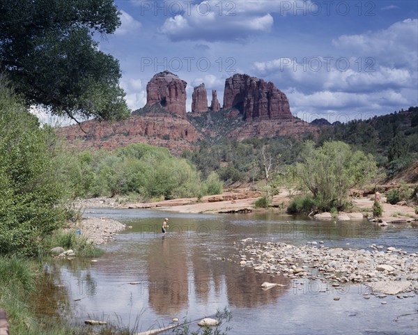 USA, Arizona, Sedona, Cathedral Rock in Oak Creek Canyon. View across river towards wind eroded rock features with a boy wading in the water.