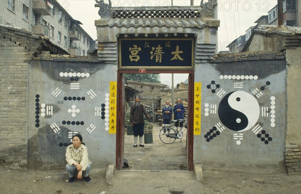CHINA, Inner Mongolia, Hohhot, Monks or priests seen through the entrance of Daoist temple with trigram and yin yang symbols on the exterior walls and person crouched outside.