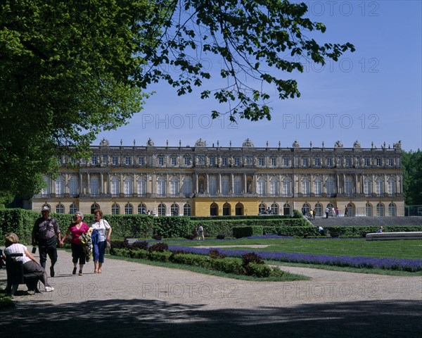 GERMANY, Upper Bavaria, Chiemgau, Chateau Herrenchiemsee. View across gardens to chateau with visitors walk on paths. Trees to the left and clear blue sky.