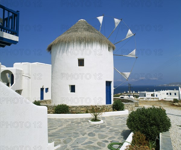 GREECE, Cyclades Islands, Mykonos, "White windmill with thatched roof, sails have flags at tips, blue door, white town buildings & coast beyond. nr.06.053579.00 "