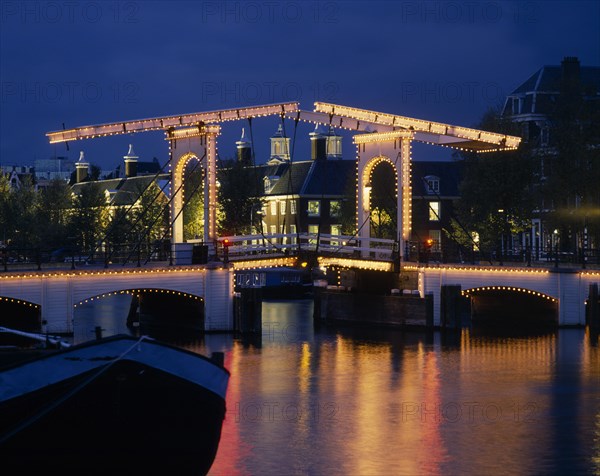HOLLAND, Noord, Amsterdam, Magere Brug the Skinny Bridge illuminated at night with a blue sky and houses in the  background
