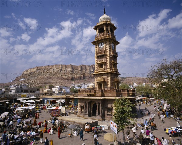 INDIA, Rajasthan, Jodphur, Sadar Market. Clock tower surrounded by stalls and traders with hillside behind.