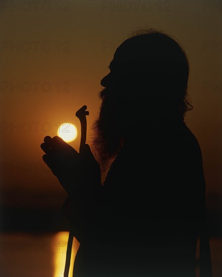 INDIA, Uttar Pradesh, Varanasi, Silhouette of Holy Man praying on the banks of the River Ganges at sunset