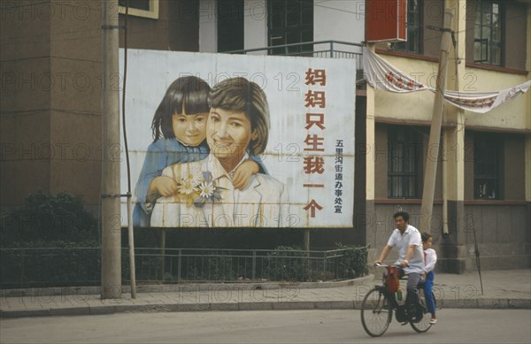CHINA, Shandong, Jinan, Man with child passenger cycling past birth control poster with image of female child.