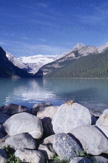 CANADA, Alberta, Lake Louise, View over boulders and clear blue water toward snow covered rocky mountains with a chipmunk sitting in the foreground