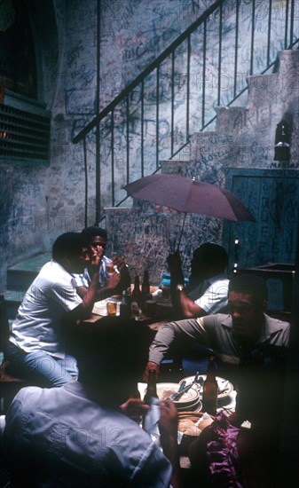 CUBA, Havana, Bodequita del Medio Bar with people sitting at tables in an alley by a stairway and one man holding an umbrella