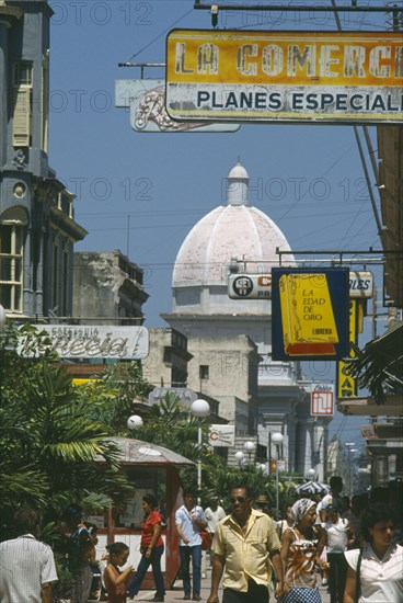 CUBA, Cienfuegos, "Busy street scene with people, hanging shop signs and dome of white building beyond."