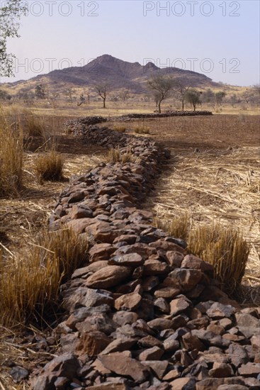 BURKINA FASO, Environment, Flooding, "Bund, low rock walls built to prevent soil erosion by flash floods. Stones are placed along the contours on gentle slopes. Sometimes the bunds are reinforced by planting tough grasses along the lines. The stones and grass encourage rain water to infiltrate the soil and reduce the amount of rain water that is lost by run-off. Any soil that has been eroded by run-off is trapped by the bund. Topsoil and organic matter (e.g. leaf litter) is deposited here.Bunds are placed 10 to 25 metres apart"